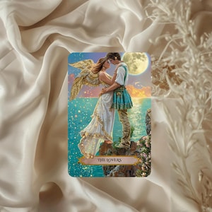 May include: A tarot card depicting a couple embracing on a cliff with a full moon and ocean backdrop. The woman wears a white dress, the man a blue and white outfit. The card is labelled "THE LOVERS" in a gold banner.