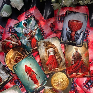 May include: A spread of tarot cards with red and gold accents. The cards feature illustrations of figures, symbols, and scenes. The cards are laid out on a dark surface with a green plant and a green stone.