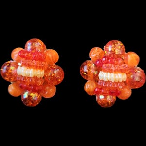 May include: A pair of orange clip-on earrings. Each earring features a cluster of orange and clear beads, with a central section of white and red beads. The earrings have a floral design and are set against a black background.