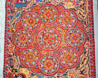 Puede incluir: Un textil vibrante, pintado a mano, con una disposición central de siete diseños circulares tipo mandala en rojo, naranja, amarillo y azul. La obra de arte está enmarcada por un borde con patrones y figuras intrincados, sobre un fondo beige.