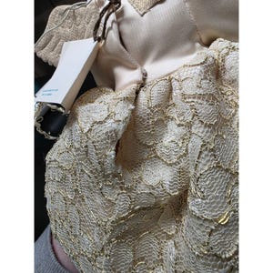 May include: A close-up of a doll wearing a cream-colored dress with gold lace overlay. The doll has a tag attached. The doll's wings are visible, made of a cream-colored material.
