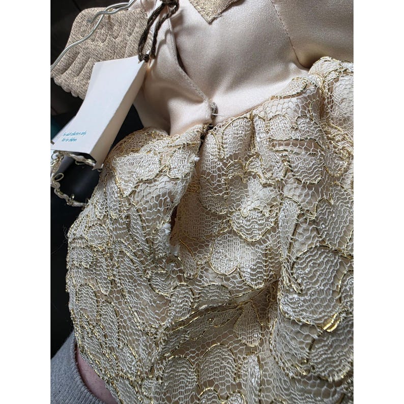 May include: A close-up of a doll wearing a cream-colored dress with gold lace overlay. The doll has a tag attached. The doll's wings are visible, made of a cream-colored material.