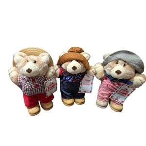 May include: Three plush teddy bears with light brown fur and tan muzzles. The left bear wears a straw hat and red plaid shirt with red pants. The center bear wears a brown hat, plaid shirt, and blue overalls. The right bear wears a gray cap and red striped overalls.