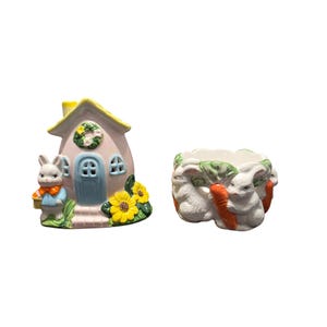 May include: Two ceramic Easter decorations. One is a pink house with a blue door, yellow roof, and a bunny figurine. The other is a white planter with bunnies and carrots.
