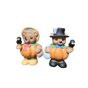 Op de afbeelding: Twee keramische berenbeeldjes in pompoenkostuums. Eén beer draagt een muts en houdt een zwarte vogel vast, de andere een hoge hoed en een vlinderdas, ook met een zwarte vogel. De beren hebben bruine vacht en staan.