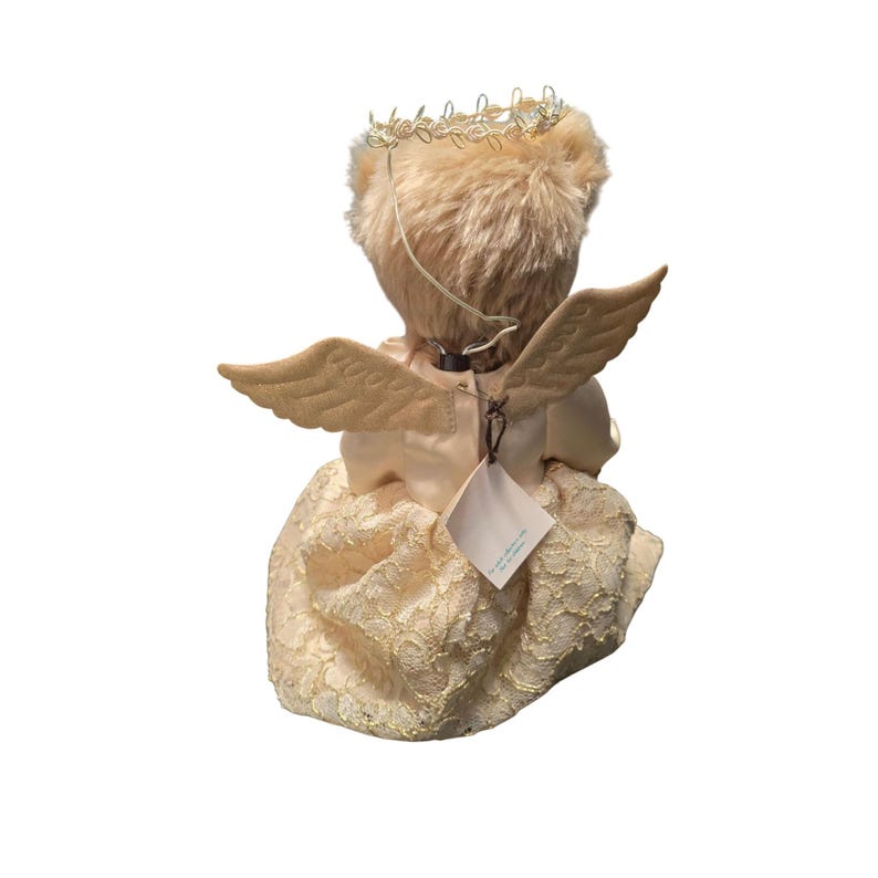 May include: A teddy bear dressed as an angel, viewed from the back. The bear has tan fur, a gold halo, and beige wings. It wears a cream-colored dress with a lace overlay. A tag hangs from the back.
