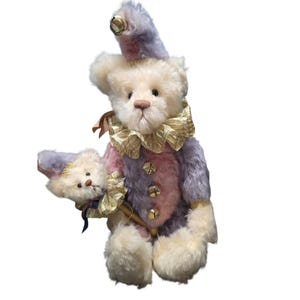May include: A plush teddy bear dressed as a jester, featuring a cream-colored face and paws, and a multi-colored body in shades of purple, pink, and cream. The bear wears a gold-trimmed collar and hat with a bell. It holds a smaller matching bear.