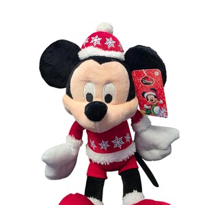 Op de afbeelding: Een Mickey Mouse pluche speelgoed in een rood kerstoutfit met witte randen en sneeuwvlokpatronen. Mickey draagt een bijpassende hoed met een witte pompon. Een Disney-label is bevestigd.