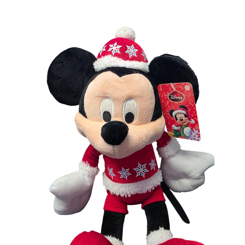 Op de afbeelding: Een Mickey Mouse pluche speelgoed in een rood kerstoutfit met witte randen en sneeuwvlokpatronen. Mickey draagt een bijpassende hoed met een witte pompon. Een Disney-label is bevestigd.