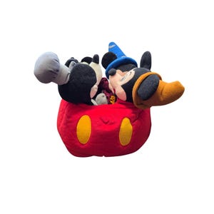 May include: A plush Mickey Mouse toy in a red, round container with yellow oval accents. Mickey Mouse is wearing a blue wizard hat and a brown and black hat. The toy is black, white, and red.