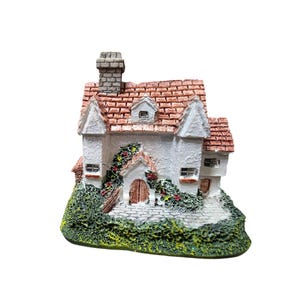 Può includere: Un cottage bianco in miniatura con tetto di tegole rosse, camino e piccole finestre. Il cottage è circondato da fogliame verde e fiori rossi, creando una scena affascinante e stravagante. La porta è marrone.
