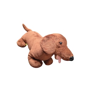 May include: A brown plush dachshund dog with a black nose and a pink tongue sticking out. The stuffed animal has floppy ears and is lying down. The toy is made of soft material.