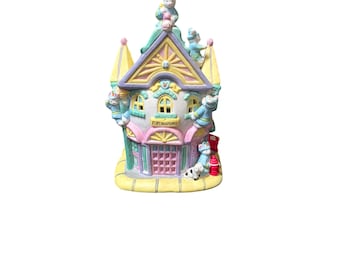 May include: A pastel-colored ceramic firehouse figurine with a yellow roof and pink door. The building has several small figures of firefighters and a Dalmatian dog. The text "FIRE HOUSE" is visible on the front.