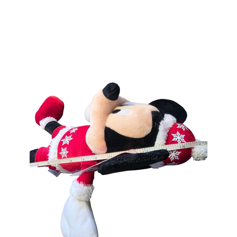 Op de afbeelding: Een pluche Mickey Mouse-speelgoed in een kerstmanoutfit. De rode outfit heeft witte randen en sneeuwvlokaccenten. Het speelgoed wordt gemeten met een meetlint. De muts en wanten zijn rood.