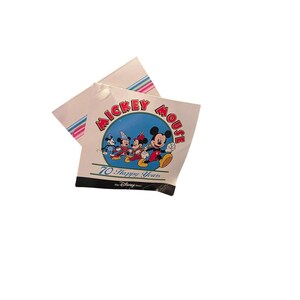 May include: A white tag featuring the words "Mickey Mouse" in a circular design with Mickey Mouse and friends. The tag also includes the text "70 Happy Years" and a colorful striped design.