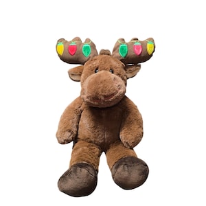 May include: A plush brown moose with embroidered antlers adorned with colorful Christmas lights. The stuffed animal has a soft, furry texture, dark brown hooves, and a friendly expression. A festive holiday decoration.