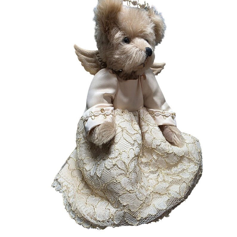 May include: A plush teddy bear dressed as an angel, wearing a cream-colored satin dress with a lace overlay. The bear has small, feathered wings, a gold crown, and a black nose. The overall design is soft and delicate.
