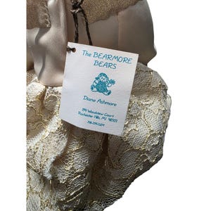 May include: A plush bear doll with a tag that reads "The BEARMORE BEARS" and "Diane Ashmore." The doll is wearing a cream-colored dress with gold lace detailing. The tag also includes an illustration of a bear.