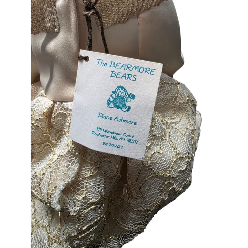 May include: A plush bear doll with a tag that reads "The BEARMORE BEARS" and "Diane Ashmore." The doll is wearing a cream-colored dress with gold lace detailing. The tag also includes an illustration of a bear.