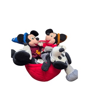 May include: Two Mickey Mouse plush toys in a red container. One Mickey wears a blue hat and a burgundy jacket. The other Mickey wears a red shirt and a brown hat. A black and white plush dog is also in the container.