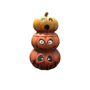 May include: A stacked pumpkin decoration with three orange gourds, each with a carved jack-o'-lantern face. The top pumpkin is a lighter shade of orange, and the stem is brown. A festive Halloween decoration.