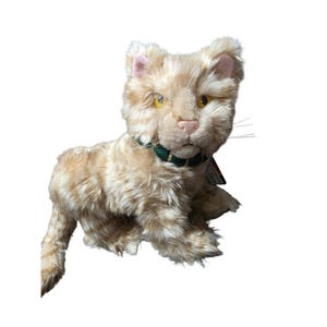 Op de afbeelding: Een pluche knuffelkat met crème en lichtoranje gestreepte vacht, gele ogen en een roze neus. Het knuffeldier heeft een donkergroene halsband met goudkleurige accenten om de nek en lange, dunne witte snorharen aan weerszijden van het gezicht.