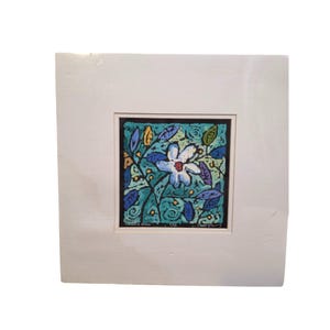 Puede incluir: Una impresión artística floral enmarcada que presenta una flor blanca con hojas azules y moradas sobre un fondo turquesa. La obra de arte está dentro de un borde negro y un passe-partout blanco, encerrada en un marco blanco. La impresión tiene el texto "Visions of Bloom".