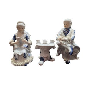 Puede incluir: Un juego de figuras de porcelana que representa a un hombre y una mujer con vestimenta del siglo XVIII, sentados con una pequeña mesa. Las figuras son blancas con detalles florales azules y adornos de encaje. La mesa tiene tazas de té y un azucarero.