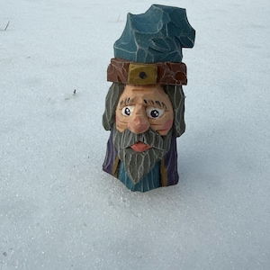 May include: A hand-carved wooden figurine of a wizard with a blue hat, brown headband, and a long gray beard. The wizard's face is detailed with a pink nose and lips. The figurine is set against a snowy background.