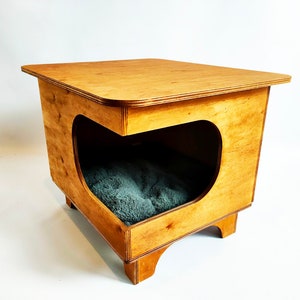 Può includere: Un letto per animali domestici in legno con finitura marrone chiaro e un cuscino grigio. Il letto ha una piattaforma rialzata e un'apertura curva per consentire all'animale di entrare.