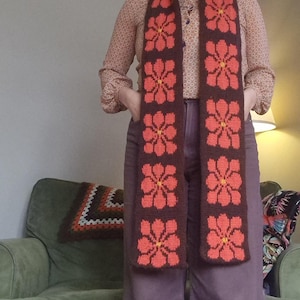 May include: A knitted scarf with a repeating floral design. The scarf has a dark brown base with large, orange flowers with yellow centres. The scarf is worn over a patterned blouse and purple trousers.