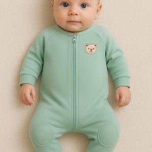 Op de afbeelding: Lichtgroene babyslaappak met een lange ritssluiting en een kleine berenafbeelding op de borst. De slaapzak heeft voetjes en knielappen. De baby heeft blauwe ogen.