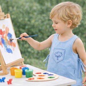 May include: A young child paints on a small wooden easel outdoors. The child wears a light blue tank top and uses a paintbrush to apply colorful paint to the canvas. Paint pots, a palette, and colored pencils are on the table.