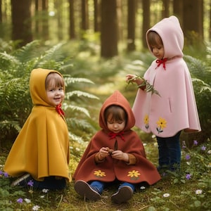 May include: Three children wearing hooded capes in a forest setting. One child wears a mustard yellow cape, another a rust-colored cape, and the third a pink cape. The capes have red bows and the pink cape has yellow flower appliques.