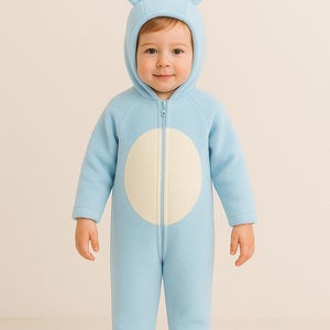 Könnte beinhalten: Ein Baby in einem hellblauen Hasenkostüm mit Kapuze und langen Ohren. Der einteilige Anzug hat einen weißen Kreis auf der Vorderseite und einen durchgehenden Reißverschluss. Das Kind lächelt und schaut in die Kamera.