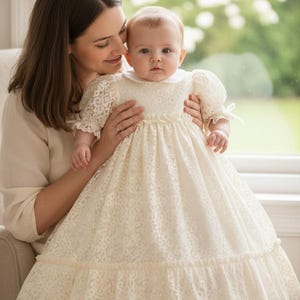 Könnte beinhalten: Ein Baby in einem cremefarbenen Spitzenkleid mit weißem Kragen und Puffärmeln. Das Kleid hat einen gerüschten Rock und wird von einer Person gehalten. Im Hintergrund ist ein Fenster mit verschwommenem Grün zu sehen.