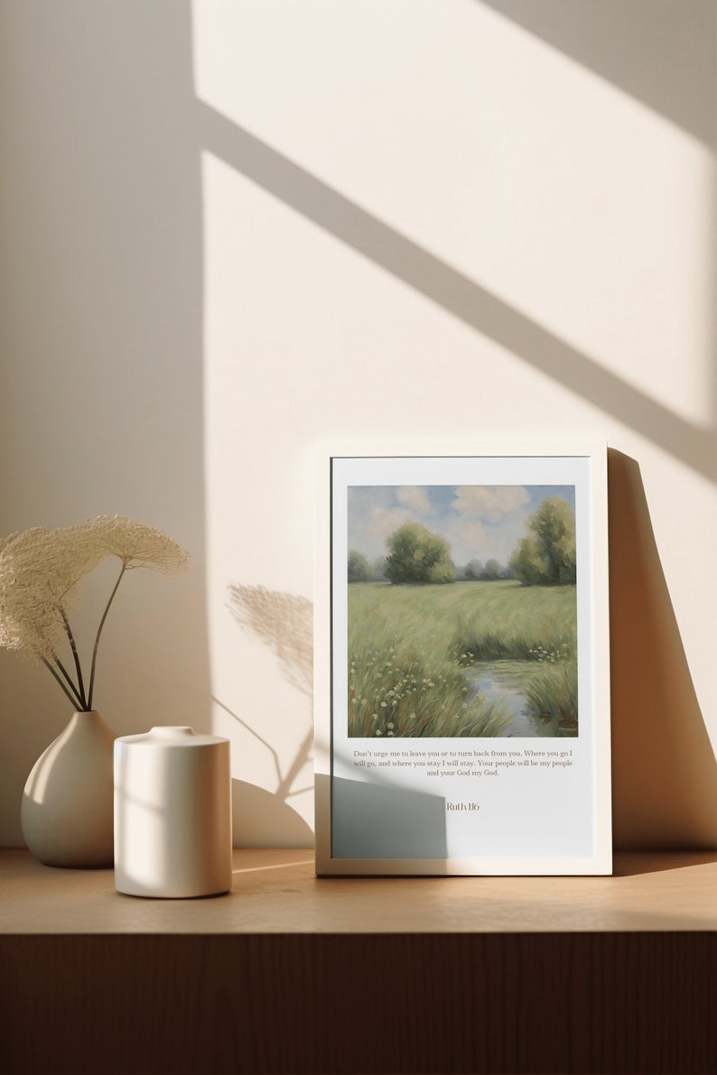 Op de afbeelding: Een ingelijste print met een schilderij van een groen veld met een beek die er doorheen loopt. De print bevat een citaat uit de Bijbel: "Dring me niet om je te verlaten of om van je terug te keren. Waar je heen gaat, ga ik heen, en waar je blijft, blijf ik. Je volk zal mijn volk zijn en je God mijn God." - Ruth 1:16.