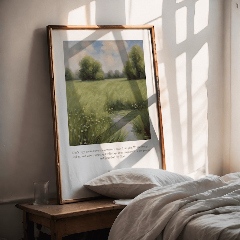 Op de afbeelding: Een ingelijste print met een schilderij van een groen veld met een beek die erdoorheen loopt. De print bevat een citaat uit de Bijbel: "Dring me niet om je te verlaten of om terug te keren van je. Waarom ik zal gaan, en waar je blijft, zal ik blijven. Je volk zal mijn volk zijn en jouw God mijn God."