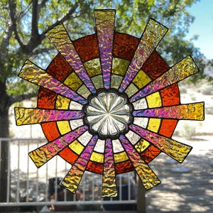 May include: A colorful stained glass suncatcher with a radial design. The center features a silver-colored, flower-like shape, surrounded by concentric rings of red, orange, yellow, and iridescent glass. The suncatcher has radiating rectangular extensions in shades of gold and purple.