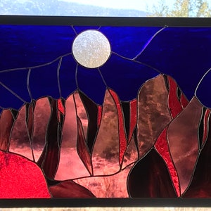 May include: A stained glass window depicting a night sky with a full moon and a mountain range in shades of red, brown, and purple.