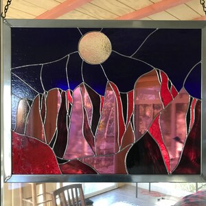 May include: A stained glass window depicting a night sky with a full moon and a mountain range in shades of red, purple, and blue.