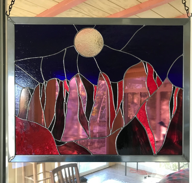 May include: A stained glass window depicting a night sky with a full moon and a mountain range in shades of red, purple, and blue.