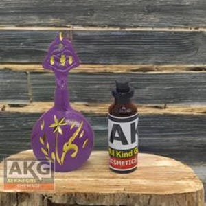 Op de afbeelding: Paarse glazen fles met een gouden bloemenontwerp en een gouden stop. De fles staat op een houten oppervlak. De fles is gelabeld met "AKG All Kind Gifts Cosmetics".