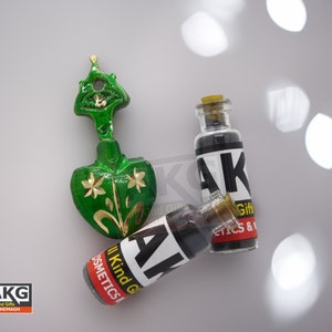 Pode incluir: Duas pequenas garrafas de vidro com líquido preto e um rótulo preto e branco que diz "AKG All Kind Gifts Cosmetics & Gifts". As garrafas estão ao lado de um ornamento de cerâmica verde em forma de coração com detalhes dourados.