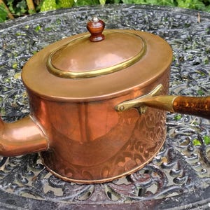 Könnte beinhalten: Eine Kupferteekanne mit einem Holzgriff und einem Messingband um den Deckel. Die Teekanne hat ein verziertes Design an der Seite.