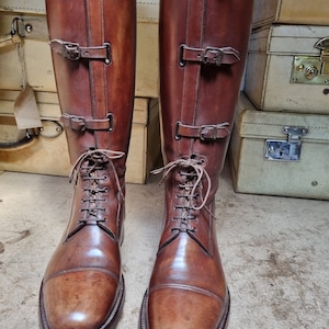 May include: A pair of brown leather riding boots with laces and buckles. The boots are polished and have a slight sheen.