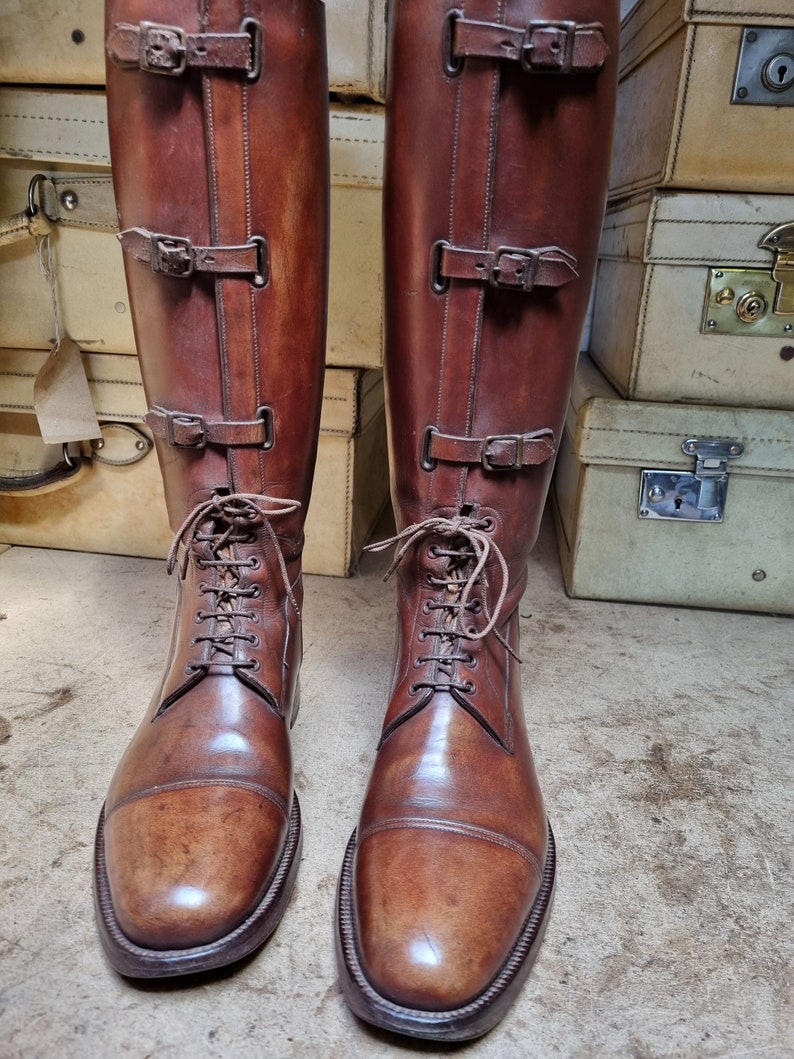 May include: A pair of brown leather riding boots with laces and buckles. The boots are polished and have a slight sheen.