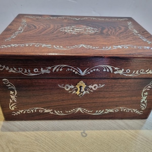 May include: A rectangular dark brown wooden box featuring intricate white mother-of-pearl floral inlay patterns across the lid and front panel. The front includes a central brass keyhole escutcheon surrounded by delicate scrolling leaf designs.