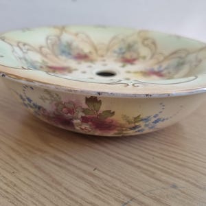 May include: A round porcelain wash basin featuring a pale cream and light green background with a floral pattern of pink, red, and blue flowers. The rim is accented with a gold-toned edge and decorative scrollwork, with a central drain hole.