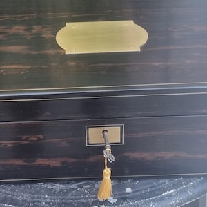 May include: A rectangular wooden box with a dark brown and black wood grain finish, featuring thin gold-colored inlay lines. The lid has a centered brass nameplate, and the front includes a brass keyhole with a metal key and a gold tassel attached.